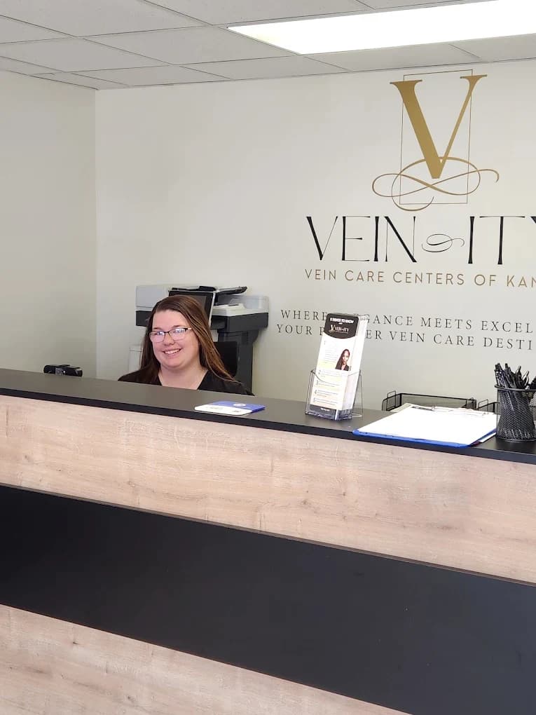 Una integrante del equipo de Vein-ity en el mostrador de recepción, bajo el logotipo de la práctica y la frase “Where elegance meets excellence”, con un exhibidor de folletos sobre el mostrador.