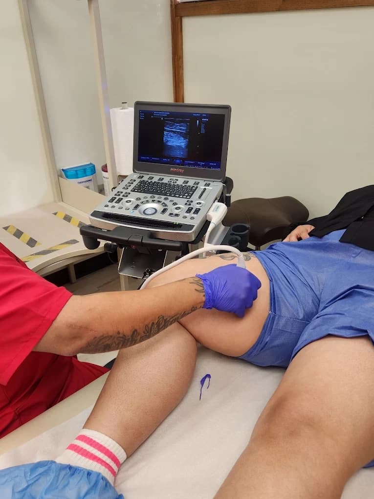 A patient lying on the examination table while a gloved clinician holds the ultrasound probe against the thigh; the machine's screen shows the scan and a marker line has been drawn on the skin.