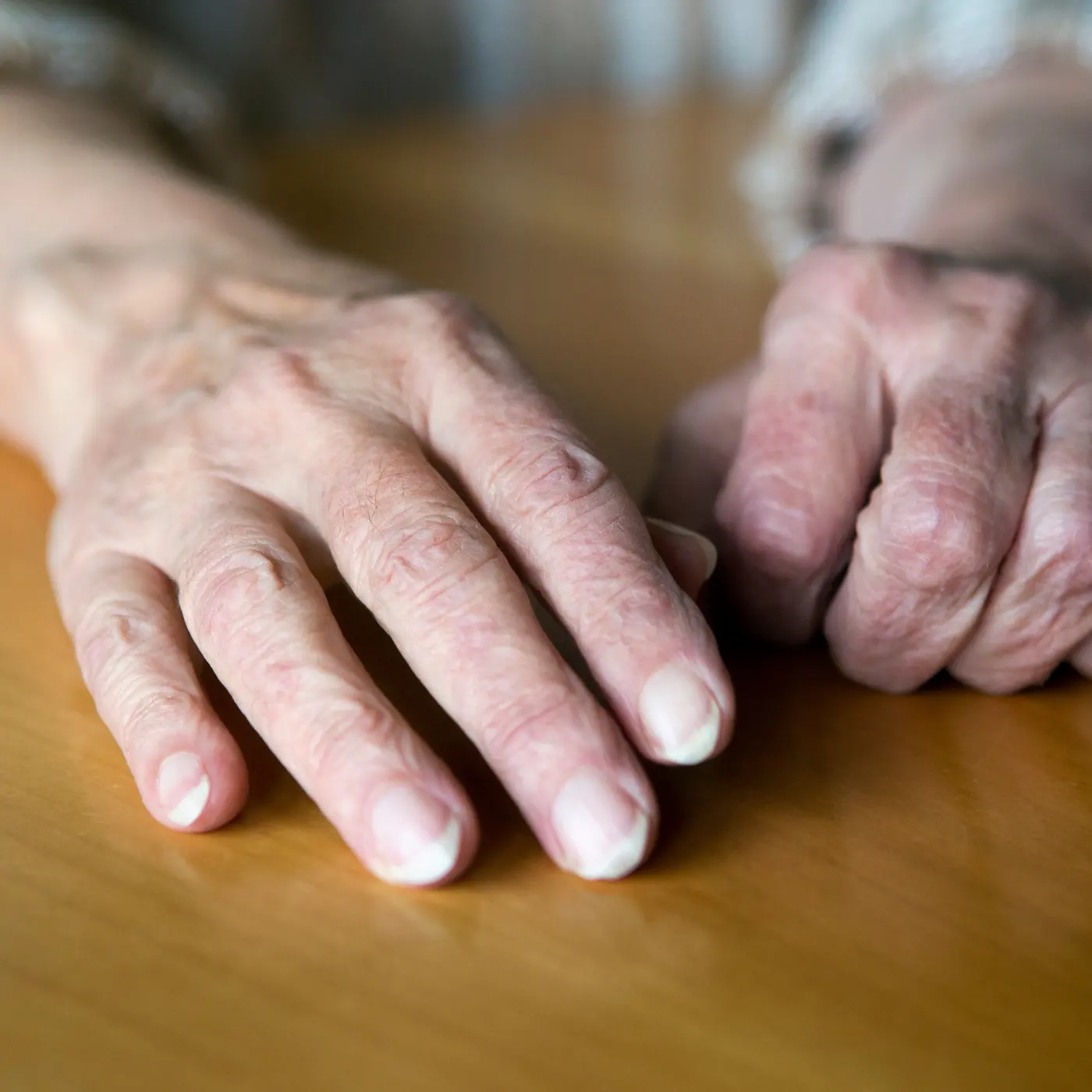 An older person's two hands resting on a wooden table, one flat and one loosely closed, with veins and tendons visible under thin skin