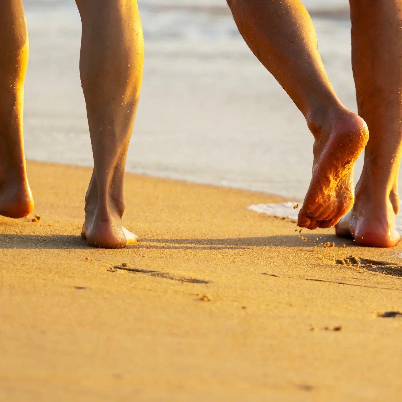 Two people walking barefoot along wet sand at the water's edge, photographed from behind at knee height in low golden light