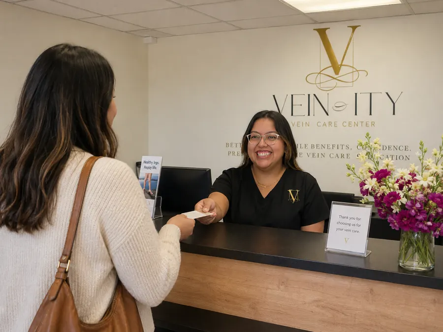 A staff member hands a card across the Vein-ity reception desk to a patient on her way out, beside a vase of flowers.