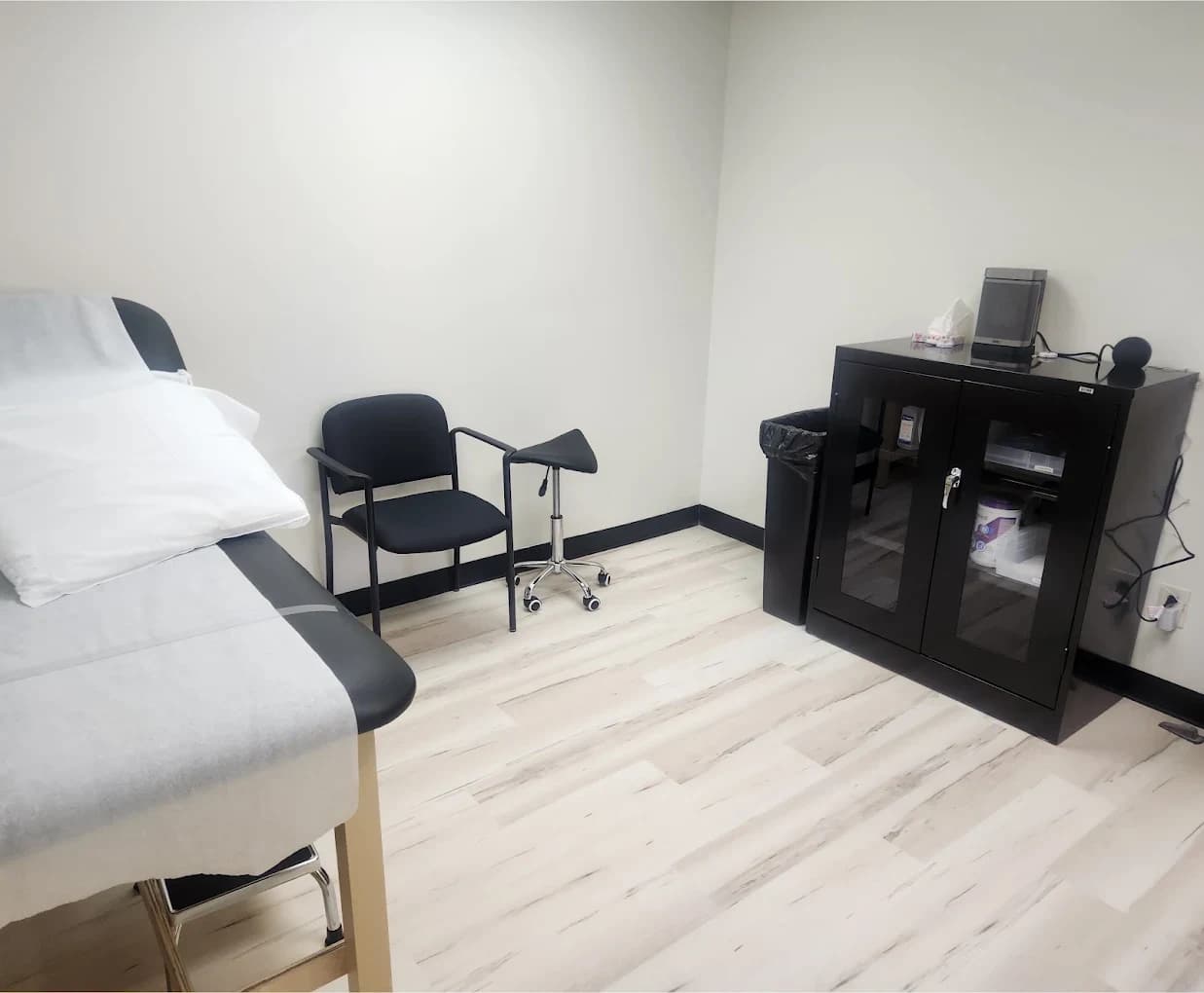 A Vein-ity examination room made up and empty: a table with a pillow and fresh linen, a chair, and a glass-fronted supply cabinet.