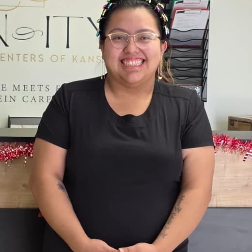 Tatiana Rodriguez smiling in glasses and black scrubs, standing at the reception desk in front of the Vein-ity wordmark.
