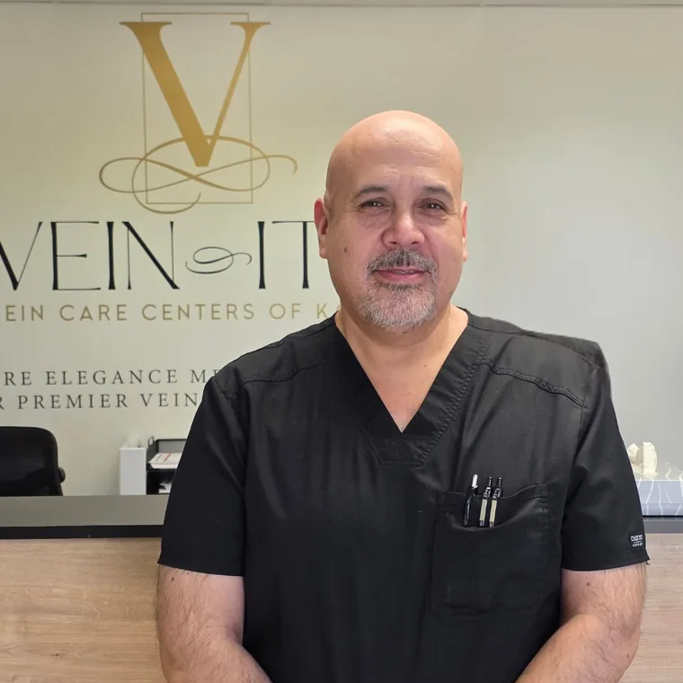 Tony Marquez con una casaca negra, de pie en el mostrador de recepción de Vein-ity bajo el logotipo de la práctica.